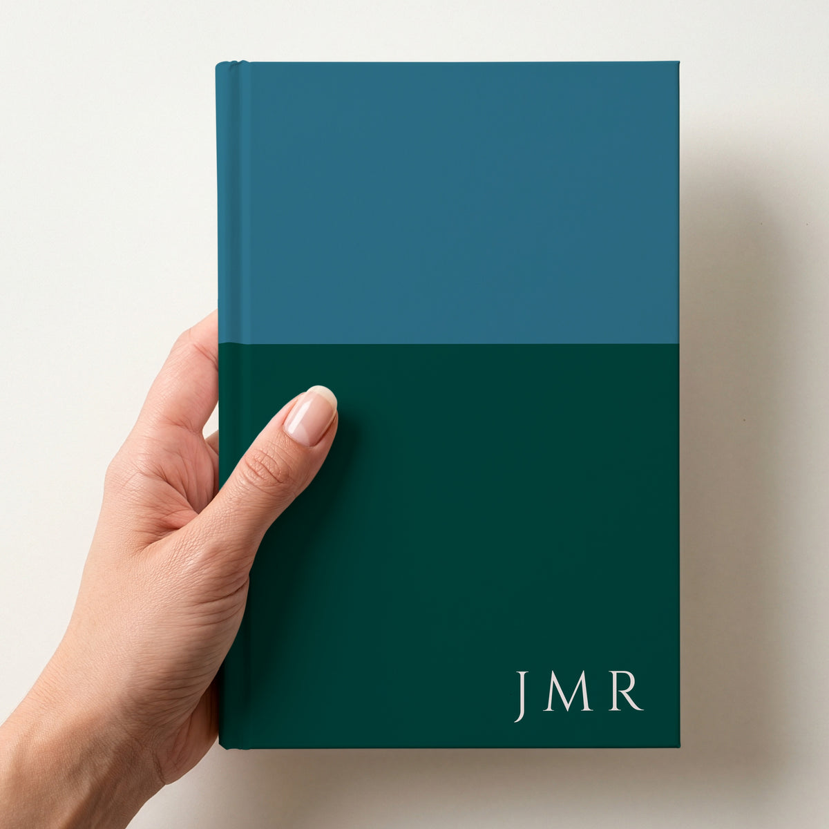 Hand holding a teal and forest two tone personalised notebook with the initials JMR in Cinzel in the lower corner