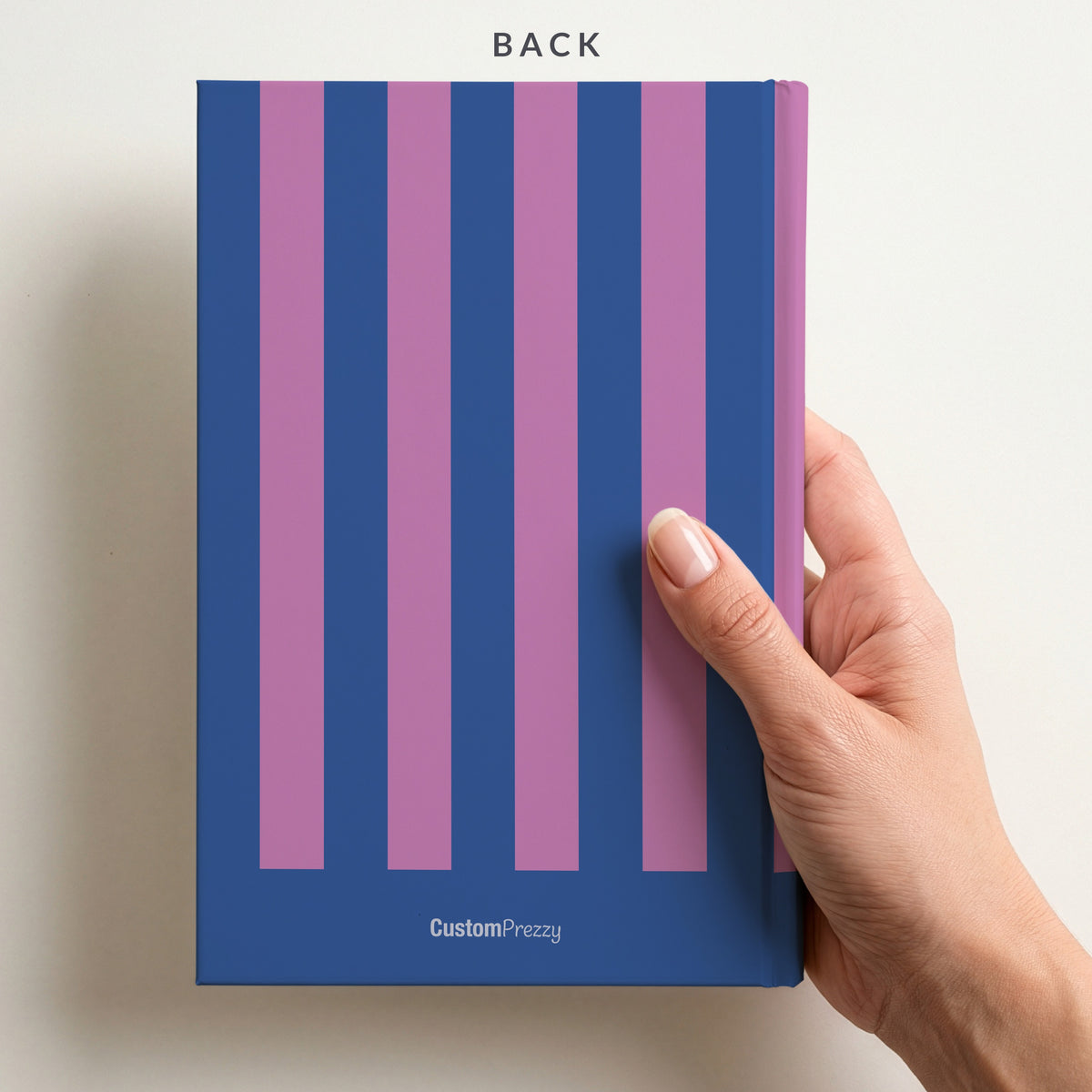 Back of the cobalt and orchid striped notebook held in a hand, with the stripes continuing round the back and the Custom Prezzy wordmark at the foot
