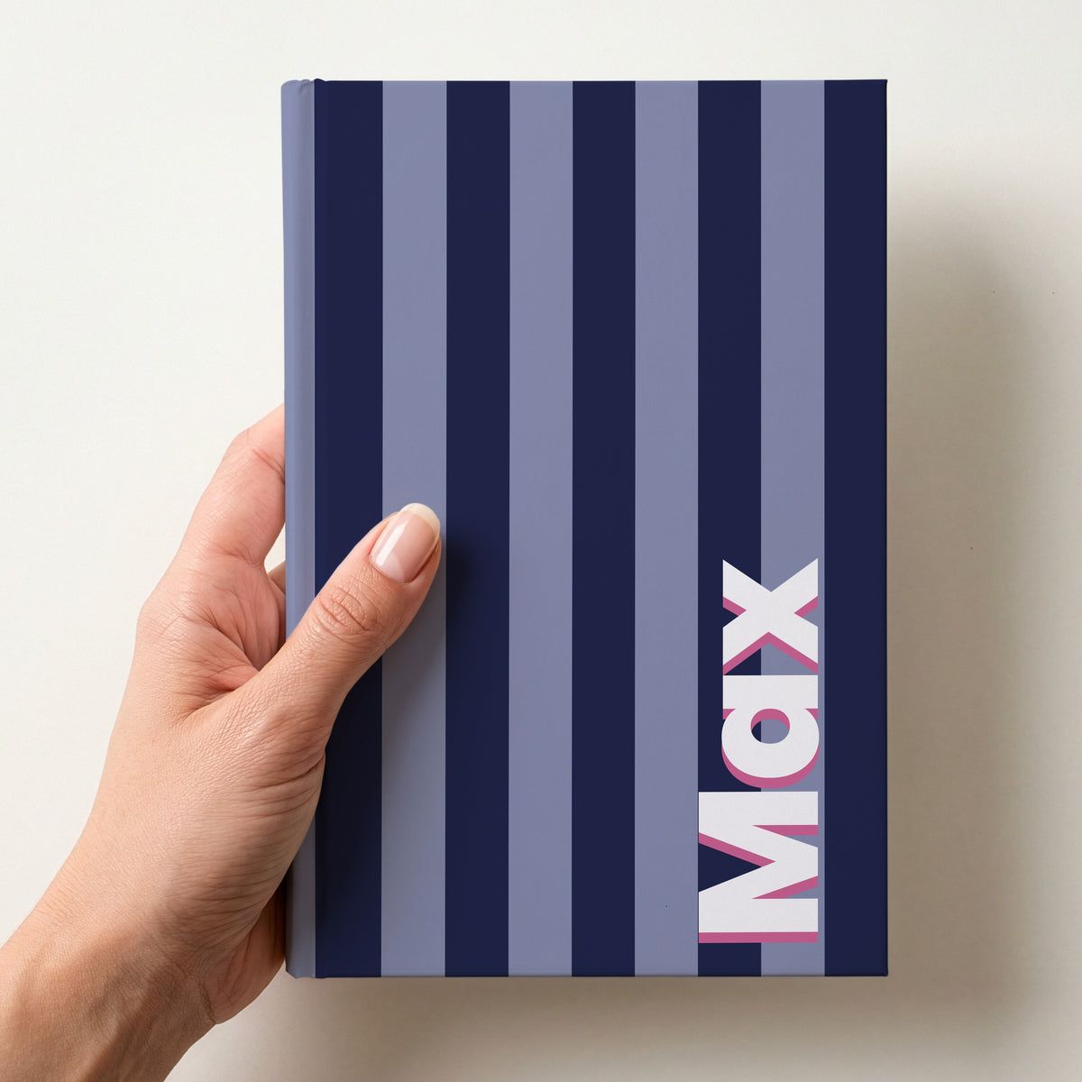 Hand holding a navy and periwinkle striped notebook with the name Max in bold block letters running up the right-hand edge