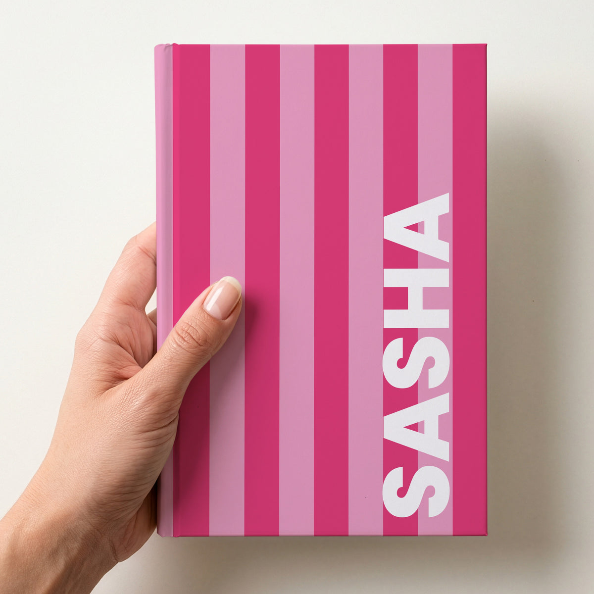 Hand holding a hot pink and candy pink striped notebook with the name SASHA in bold block letters running up the right-hand edge