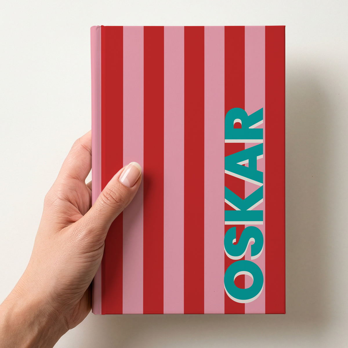 Hand holding a red and pink striped notebook with the name OSKAR in bold block letters running up the right-hand edge