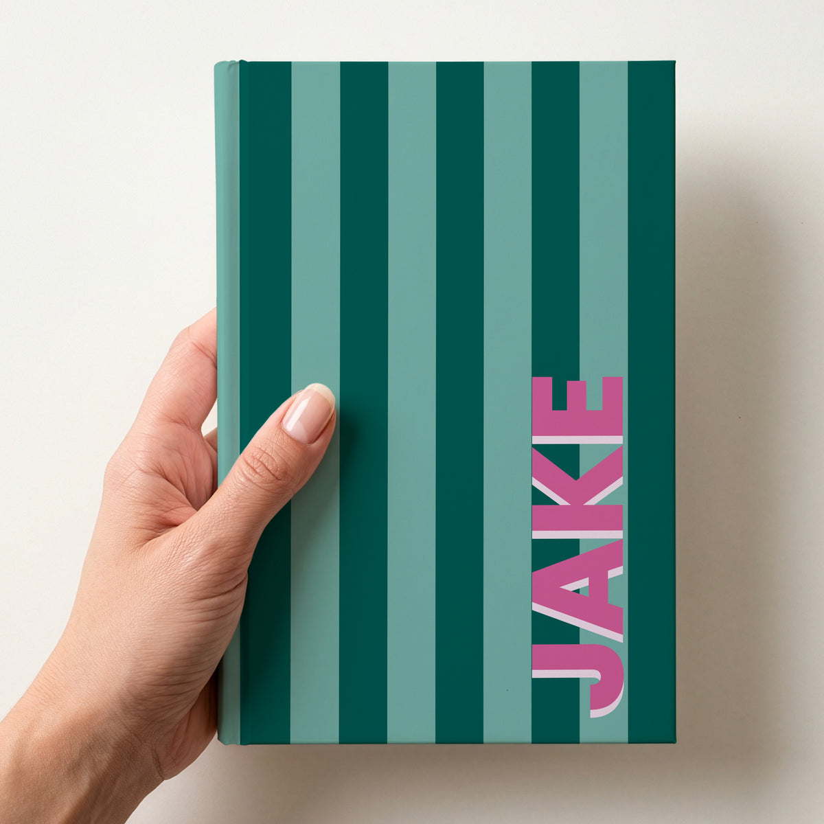 Hand holding a bottle green and sea green striped notebook with the name JAKE in bold block letters running up the right-hand edge