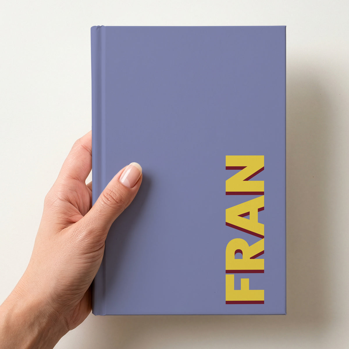 Hand holding a periwinkle blue notebook with the name FRAN in bold block letters running up the right-hand edge