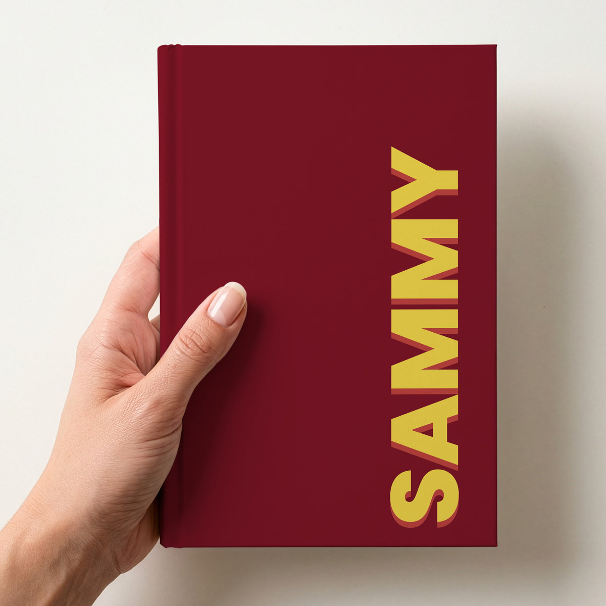 Hand holding a burgundy notebook with the name SAMMY in bold block letters running up the right-hand edge