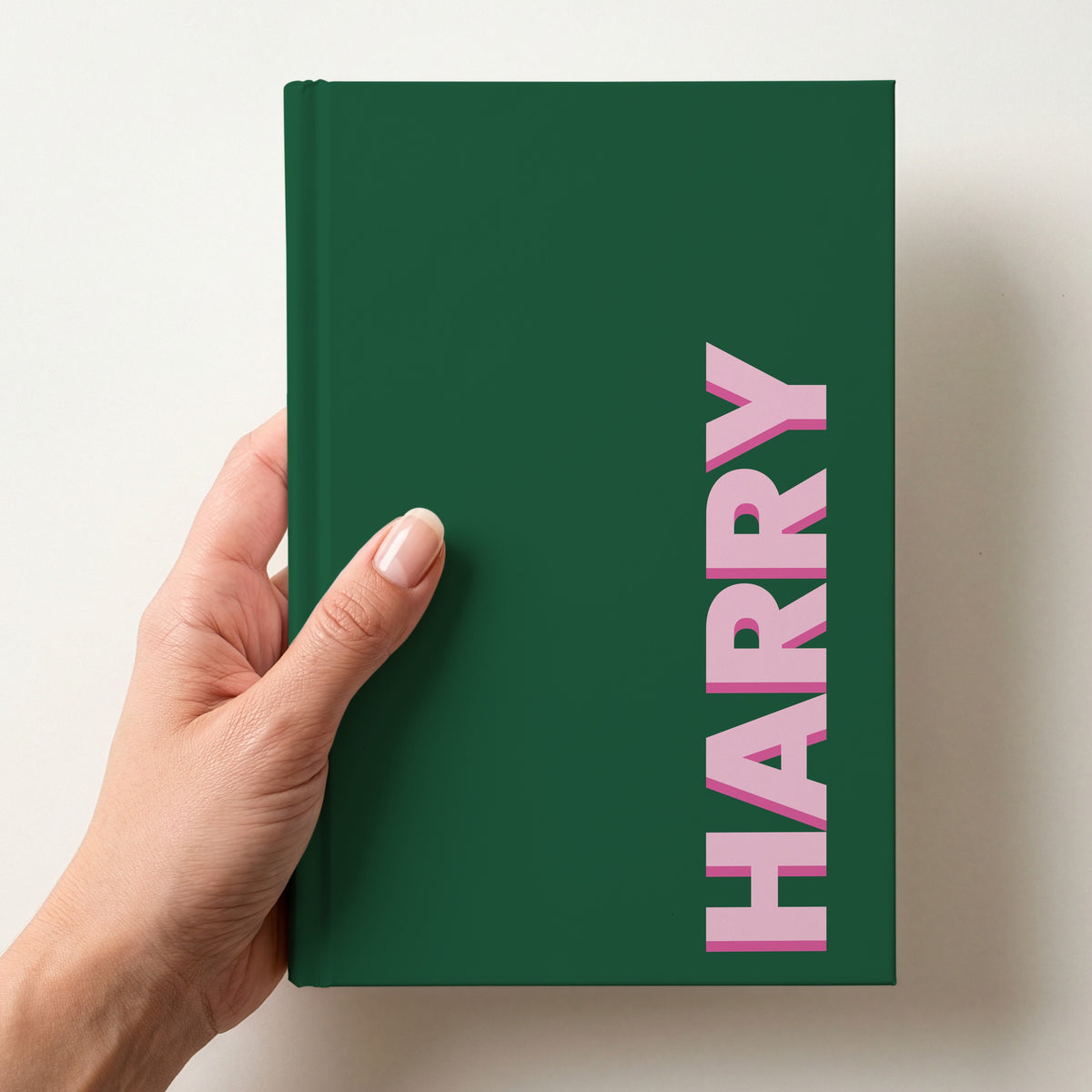 Hand holding a forest green notebook with the name HARRY in bold block letters running up the right-hand edge