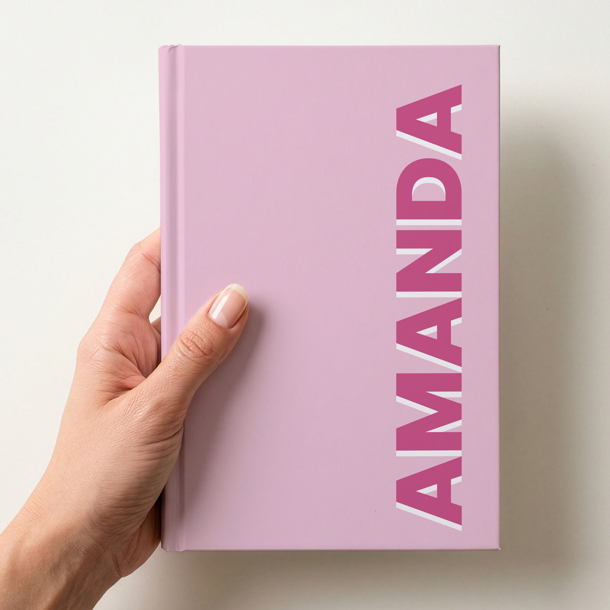 Hand holding a light pink notebook with the name AMANDA in bold block letters running up the right-hand edge