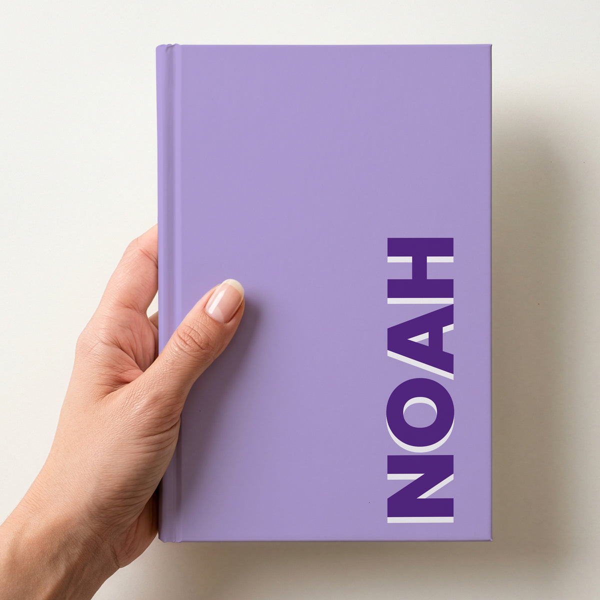 Hand holding a lilac notebook with the name NOAH in bold block letters running up the right-hand edge