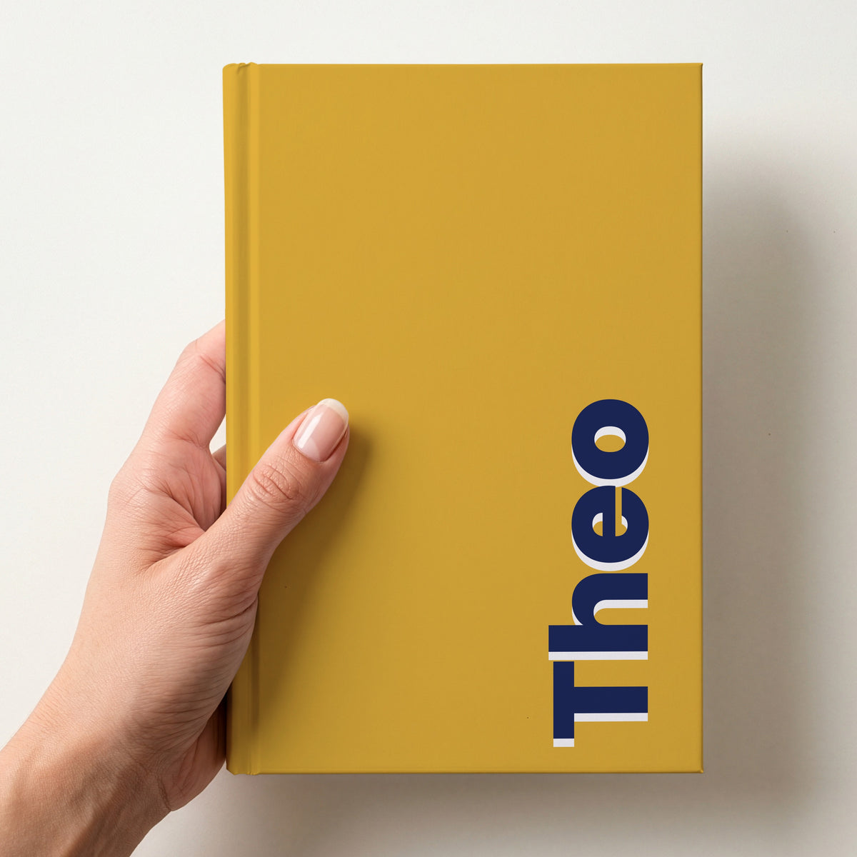 Hand holding a mustard notebook with the name Theo in bold block letters running up the right-hand edge