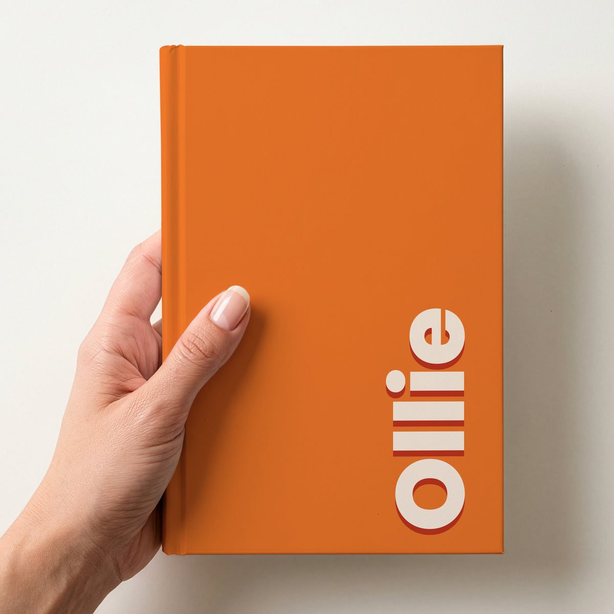 Hand holding a orange notebook with the name Ollie in bold block letters running up the right-hand edge