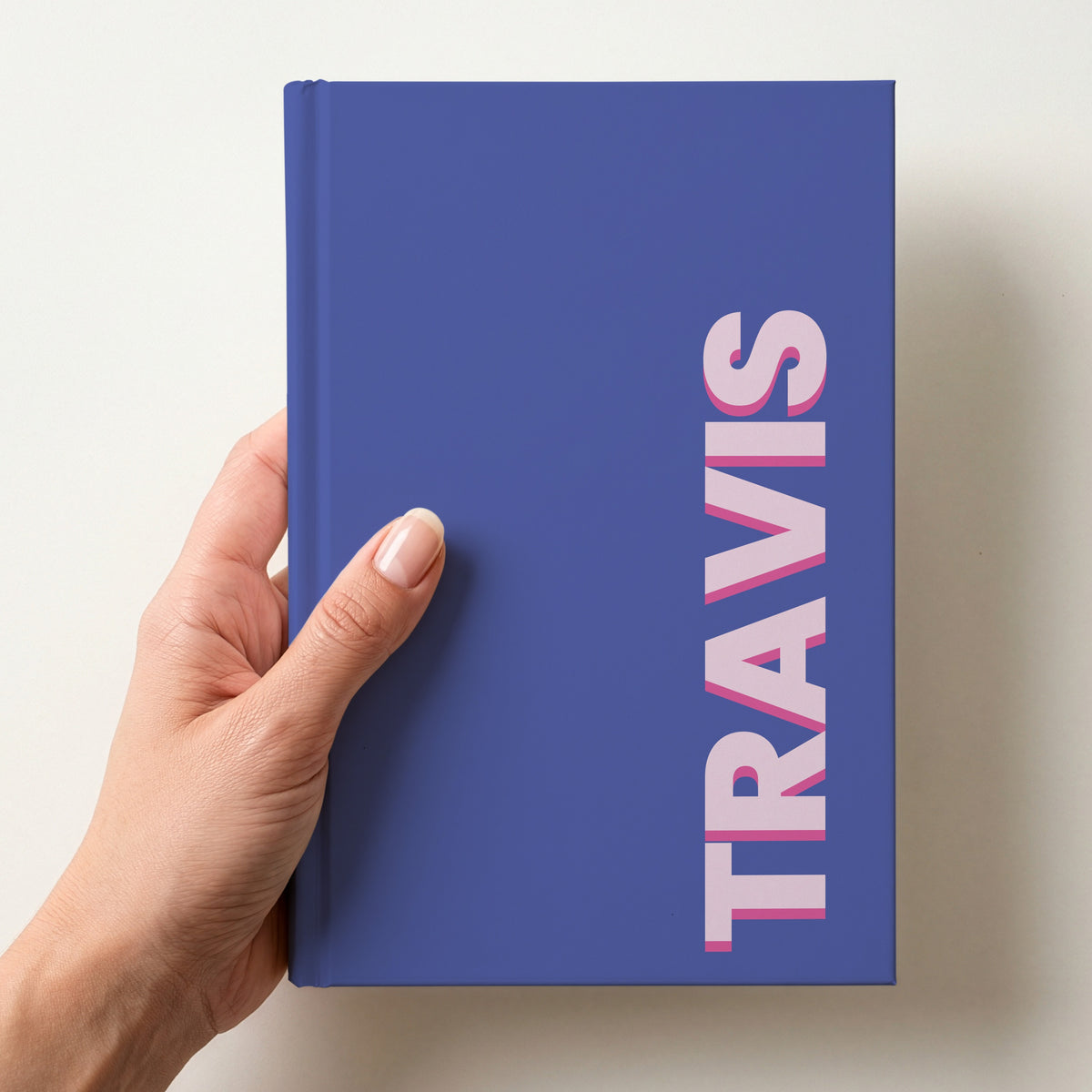 Hand holding a purple notebook with the name TRAVIS in bold block letters running up the right-hand edge