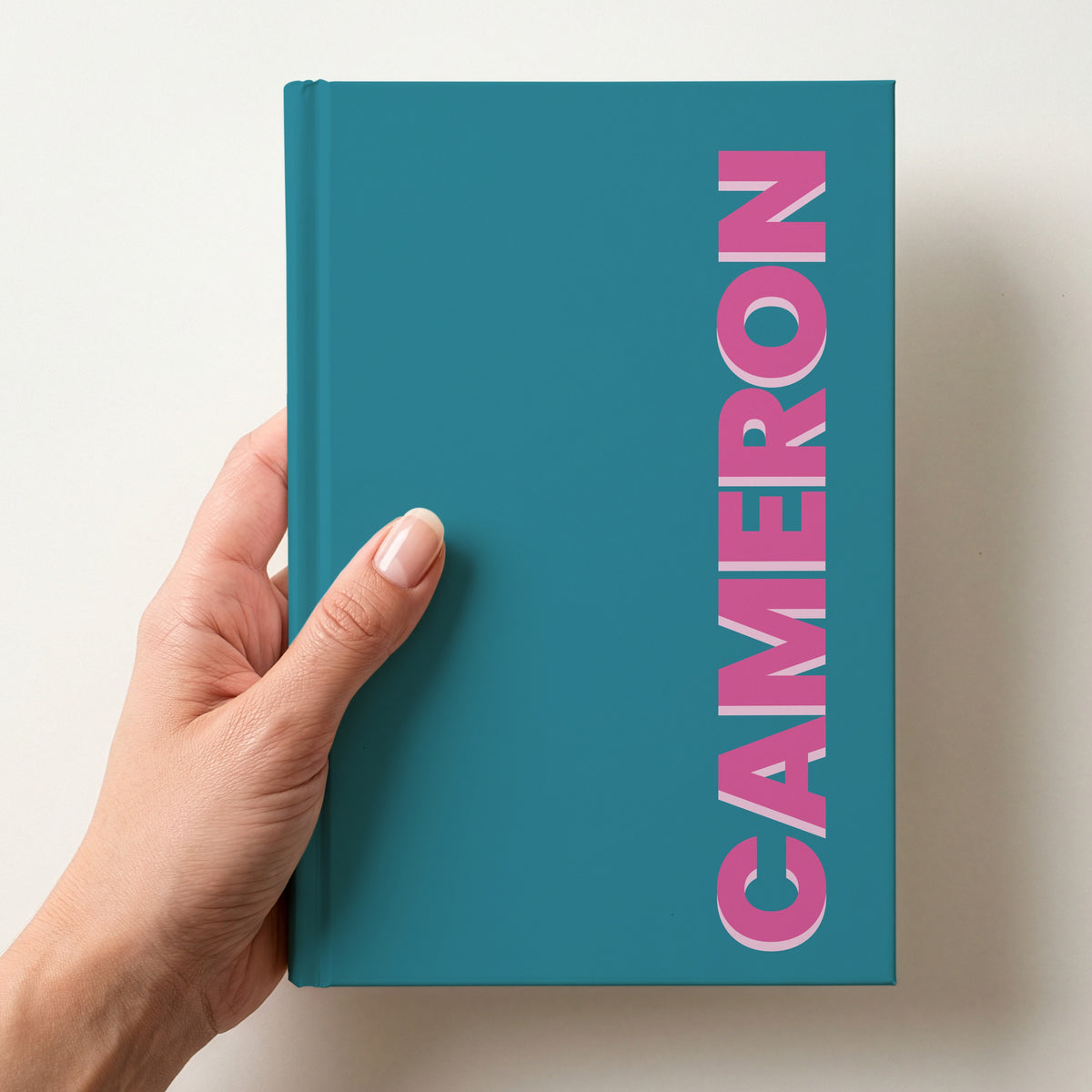 Hand holding a teal notebook with the name CAMERON in bold block letters running up the right-hand edge