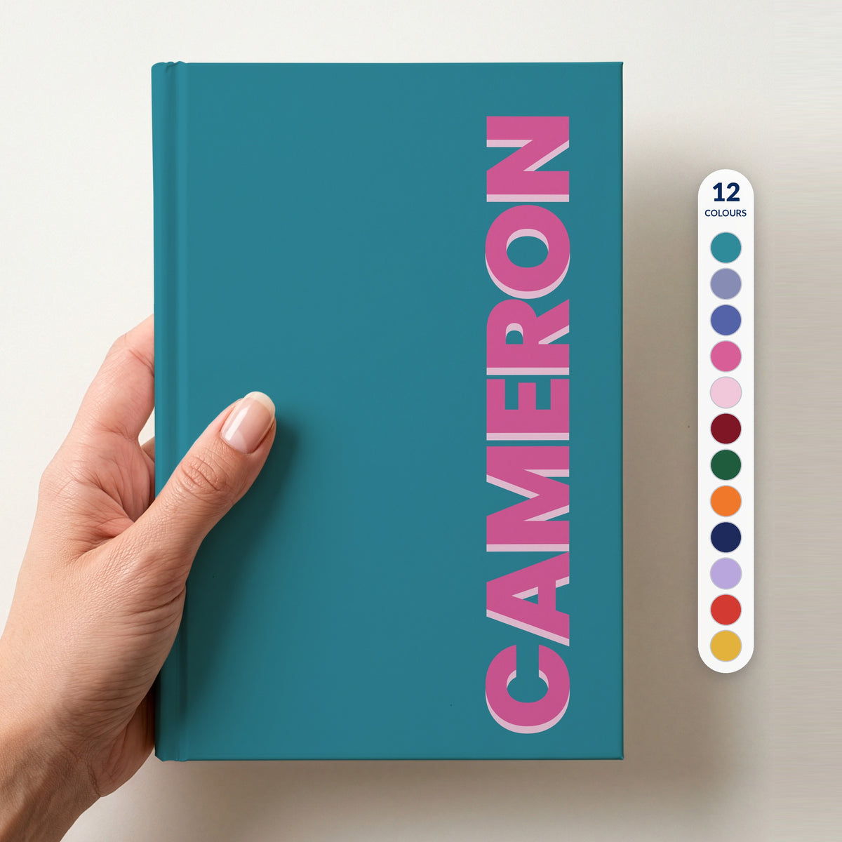 Hand holding a teal notebook with the name CAMERON in bold block letters running up the right-hand edge, with a column of 12 colour swatches beside it showing all 12 colours