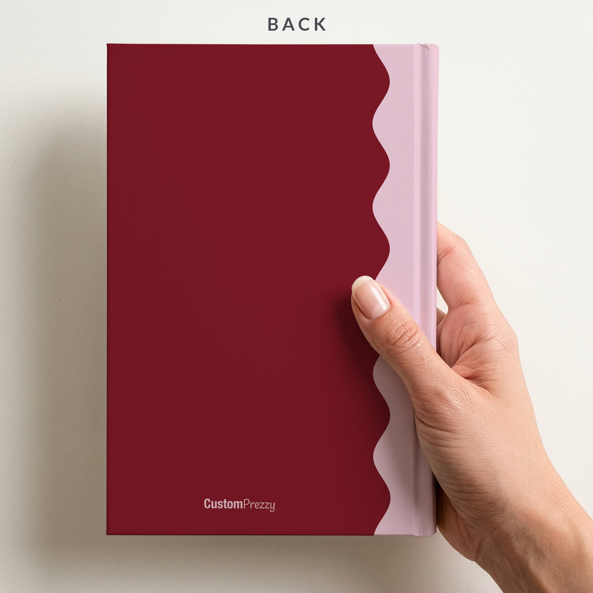 Back of the burgundy and blush scalloped monogram notebook held in a hand, with the Custom Prezzy wordmark at the foot