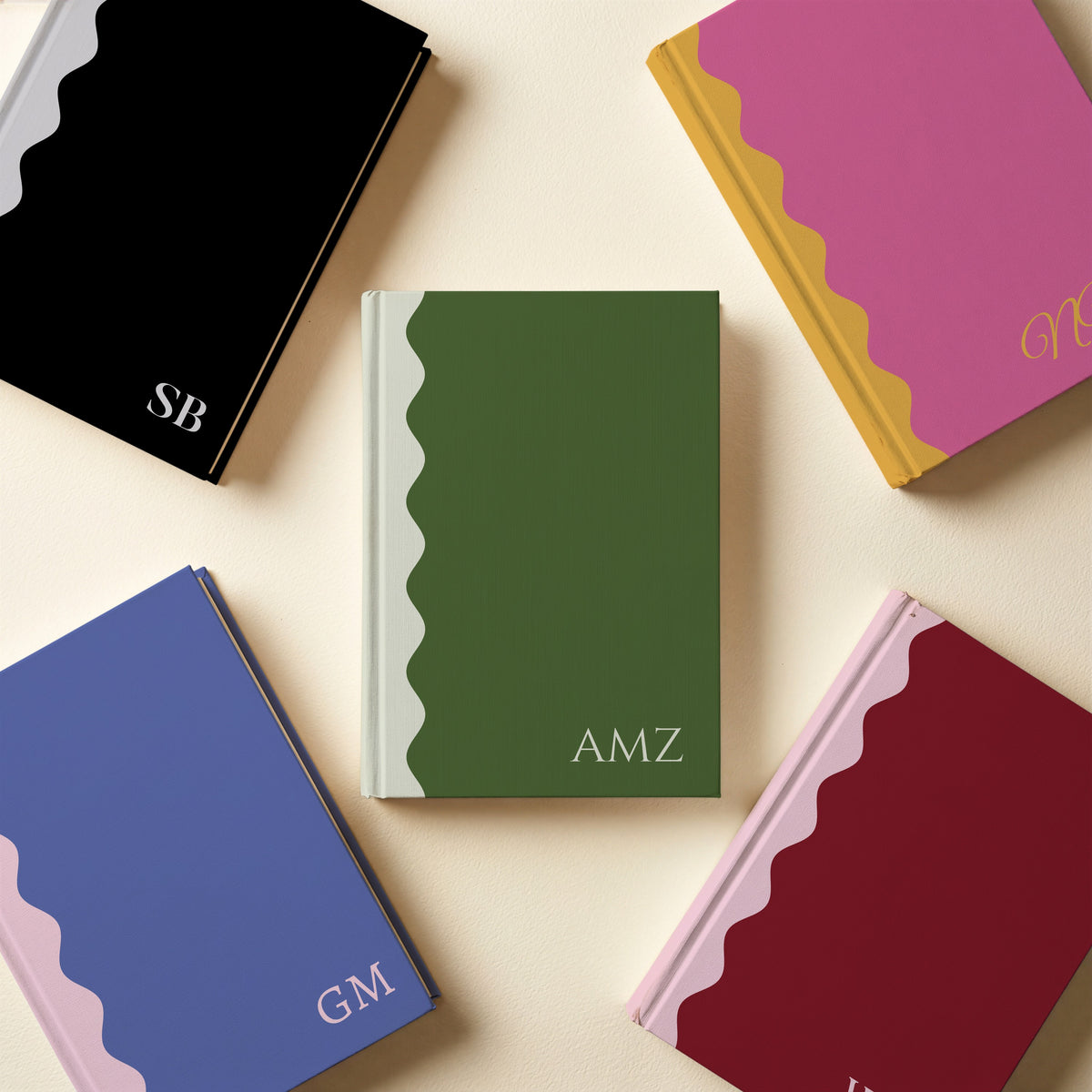 Five scalloped monogram notebooks laid flat in pink and mustard, black and grey, purple and blush, burgundy and blush, green and cream