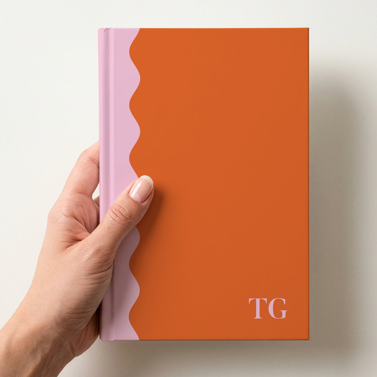 Hand holding a orange and pink scalloped colour block notebook with the initials TG in Butler in the lower corner