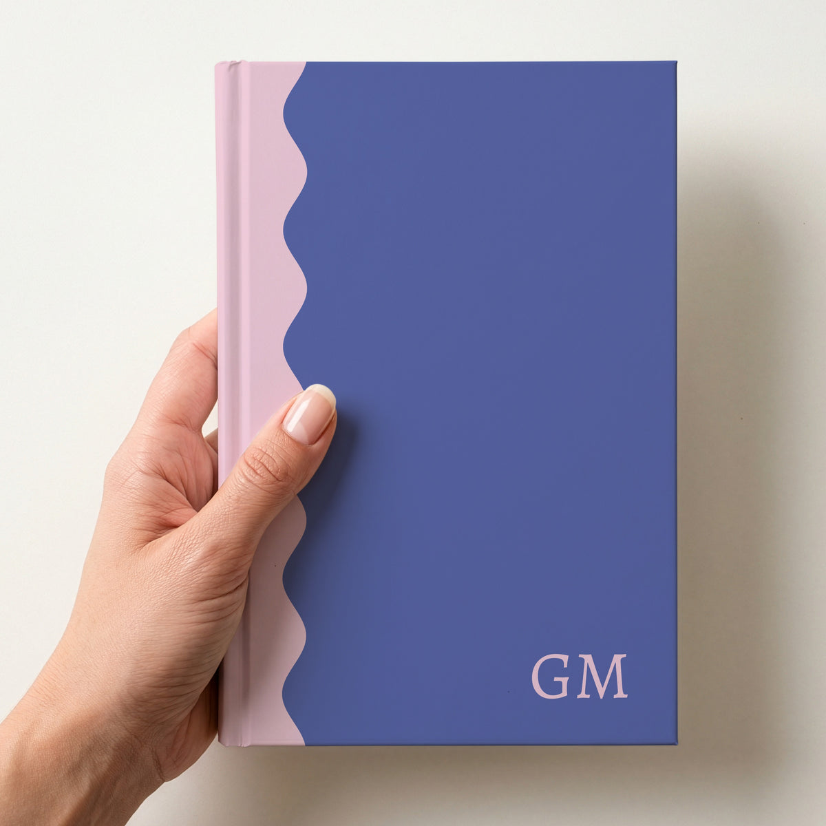 Hand holding a purple and blush scalloped colour block notebook with the initials GM in Alegreya in the lower corner