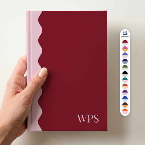 Hand holding a burgundy and blush scalloped colour block notebook with the initials WPS in Playfair Display in the lower corner, with a column of 12 colour swatches beside it showing all 12 colours