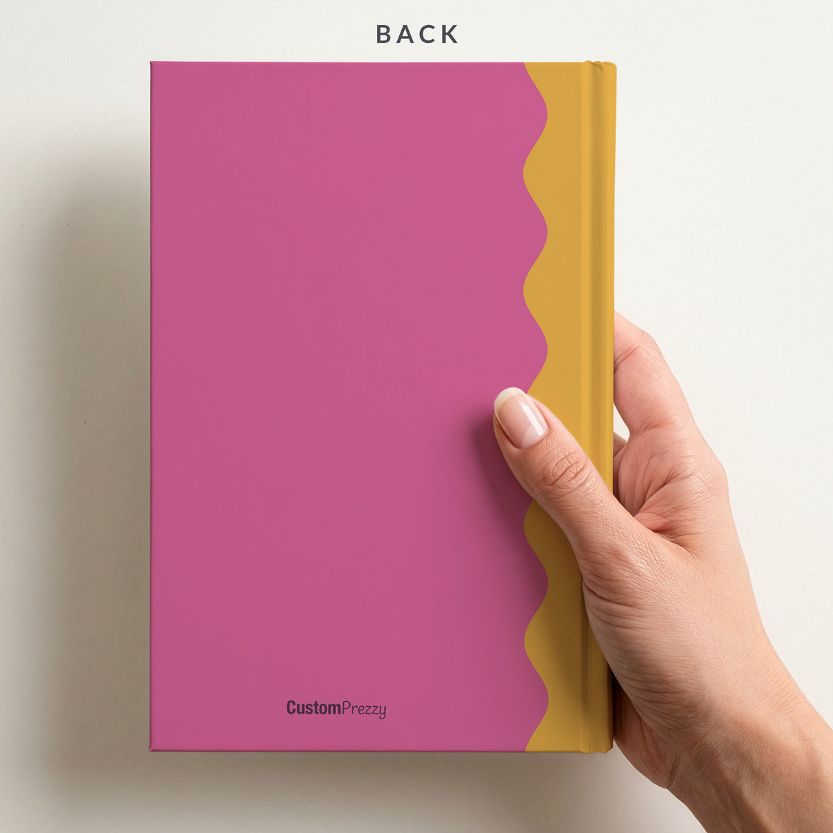 Back of the pink and mustard scalloped name notebook held in a hand, with the Custom Prezzy wordmark at the foot