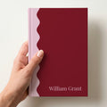 Hand holding a burgundy and blush scalloped colour block notebook with the name William Grant in Playfair Display in the lower corner