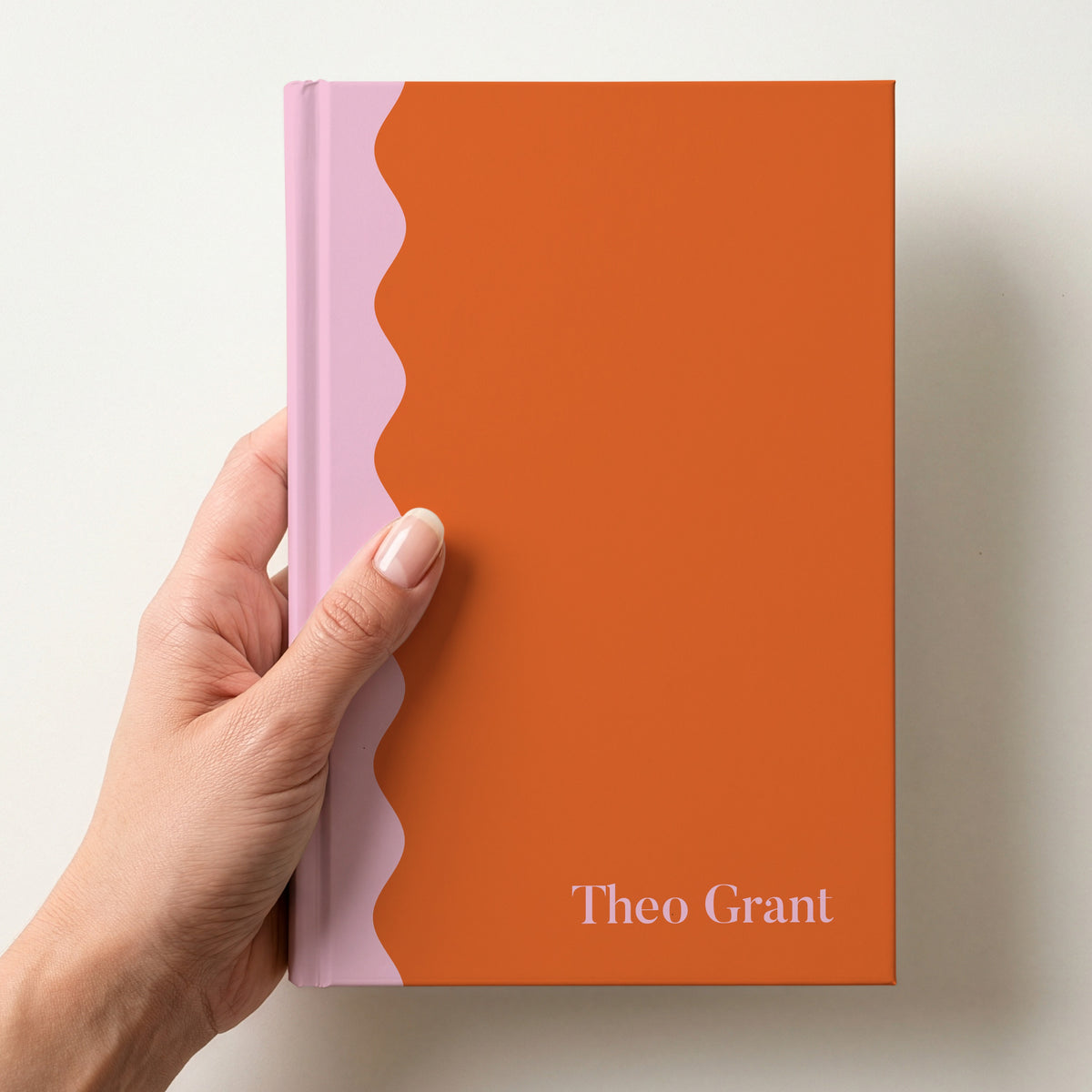 Hand holding a orange and pink scalloped colour block notebook with the name Theo Grant in Butler in the lower corner