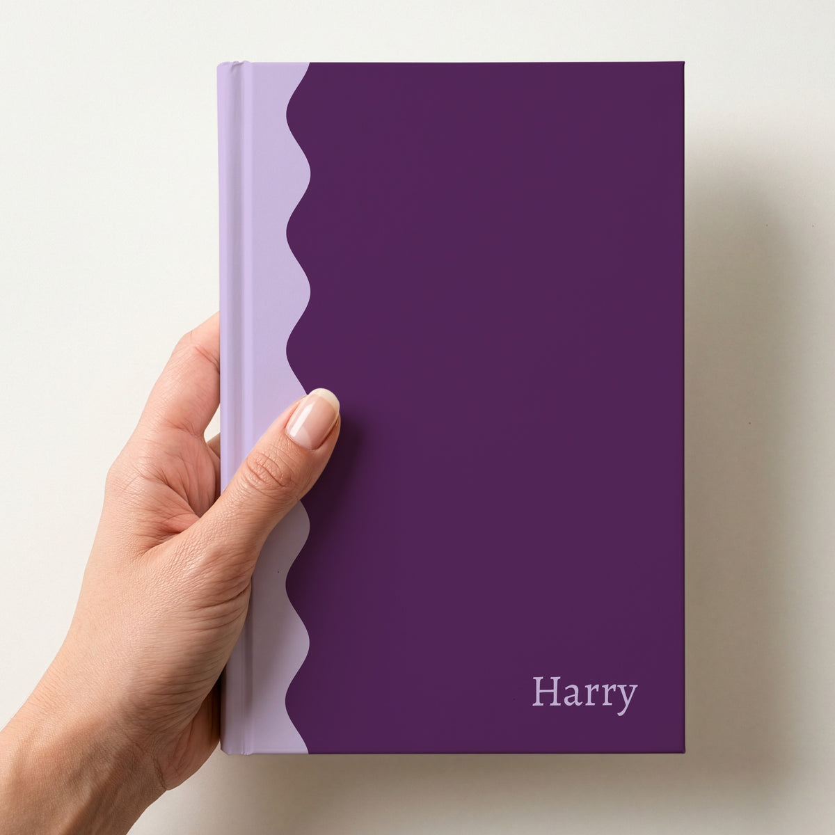 Hand holding a plum and lilac scalloped colour block notebook with the name Harry in Alegreya in the lower corner
