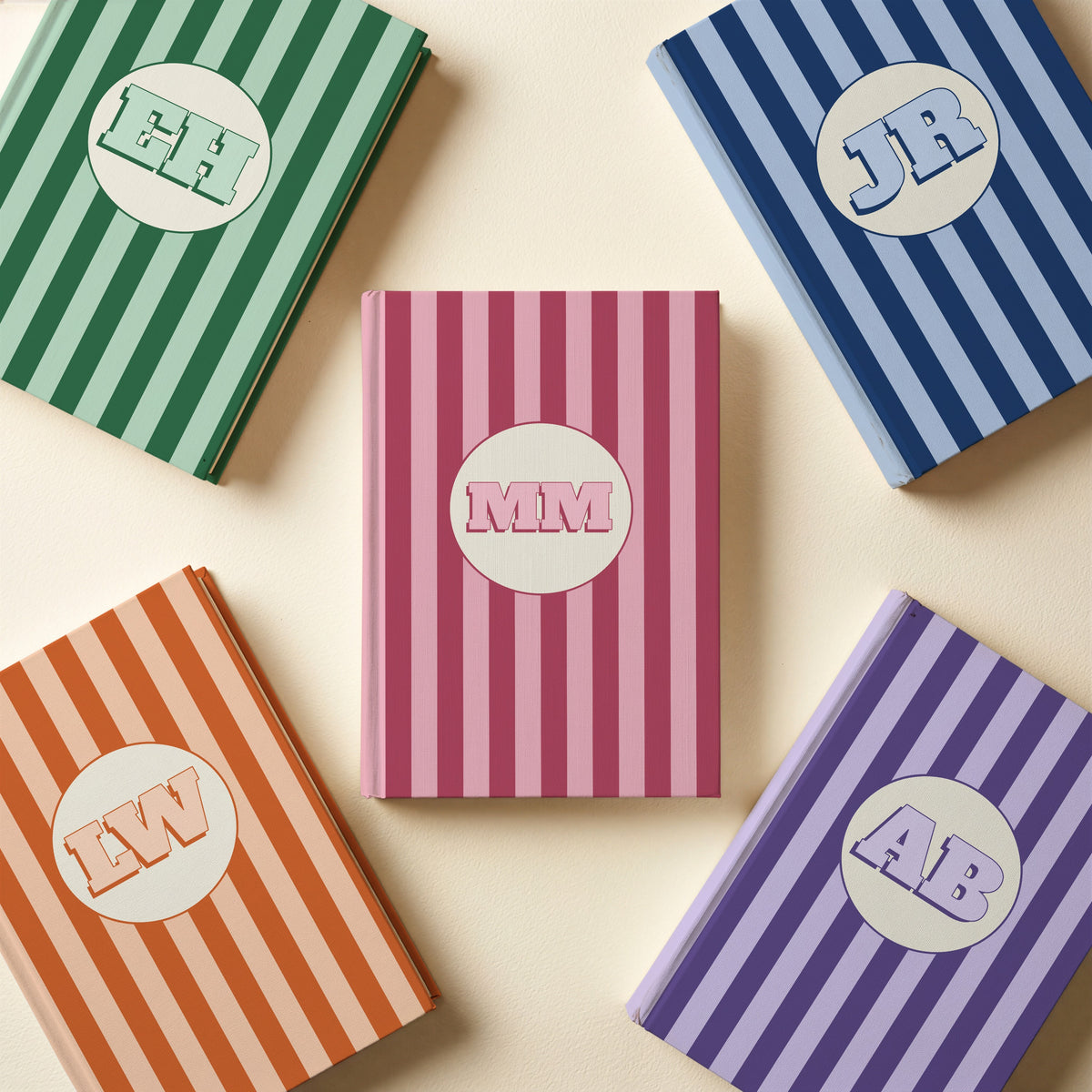 Five personalised striped monogram notebooks in pink, green, navy, orange and purple on a cream surface