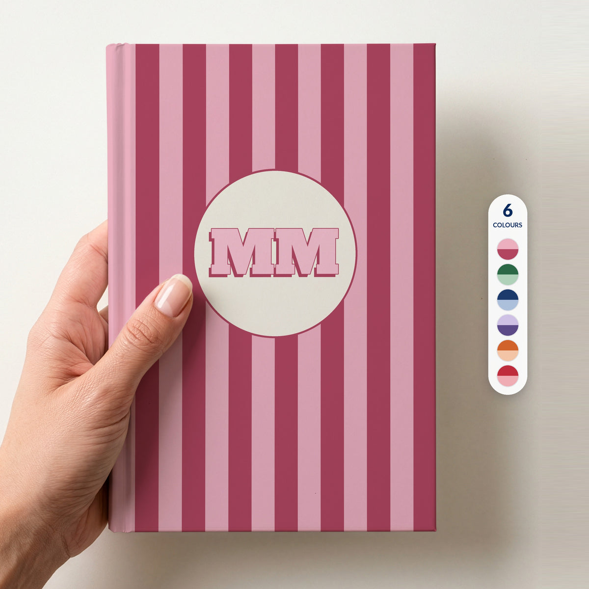 Hand holding a personalised pink striped hardcover notebook with retro initials MM in a cream circle, beside a column of 6 colour swatches showing all 6 colourways