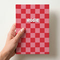 Hand holding a personalised red and pink checkerboard hardcover notebook with the name Rosie in puffy white letters