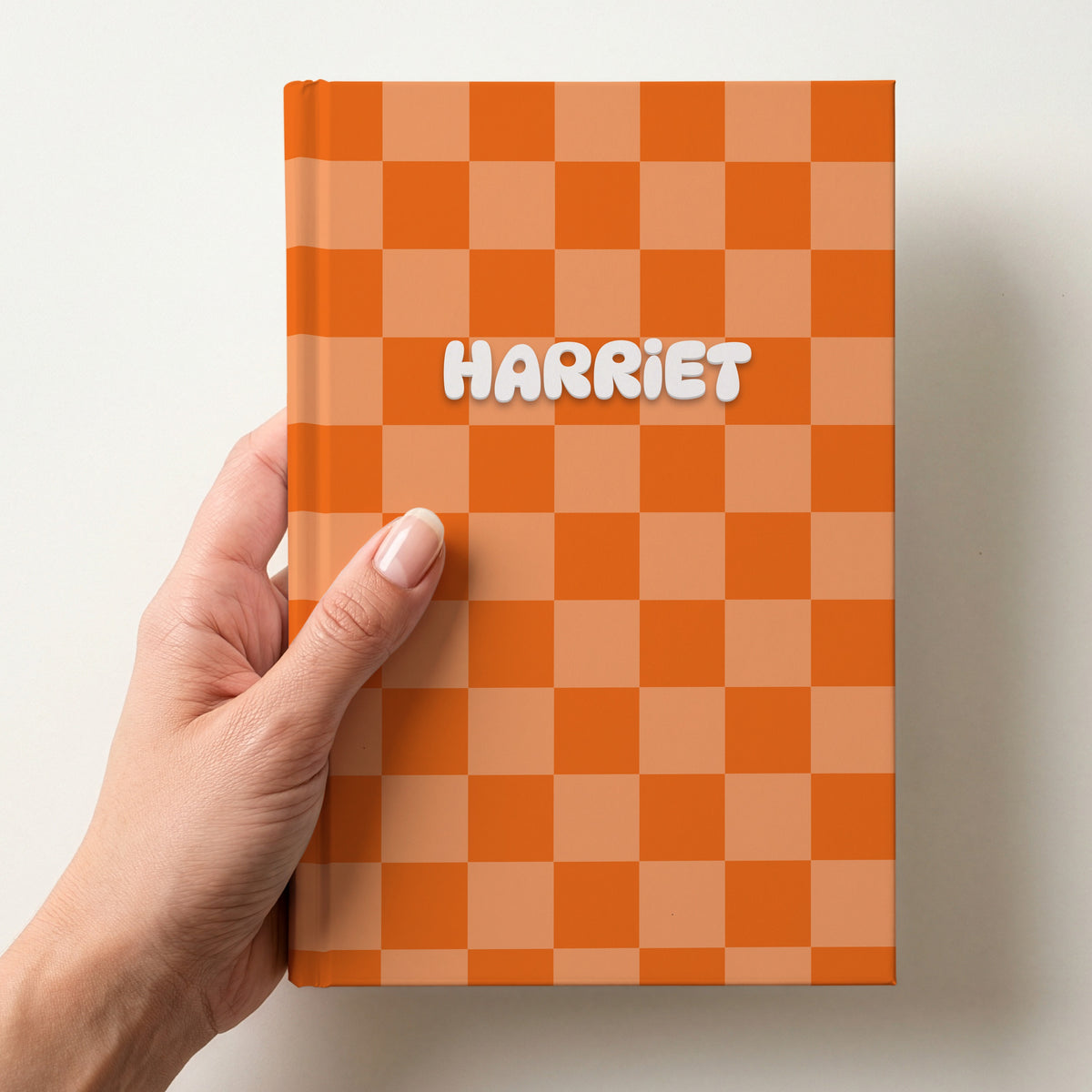 Hand holding a personalised tangerine checkerboard notebook with the name Harriet