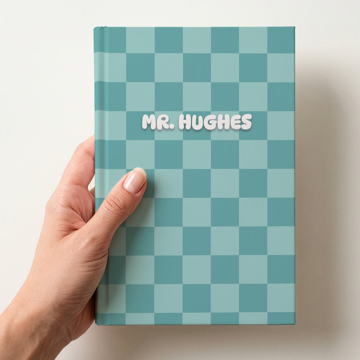 Hand holding a personalised teal checkerboard notebook with the name Mr. Hughes in puffy white letters