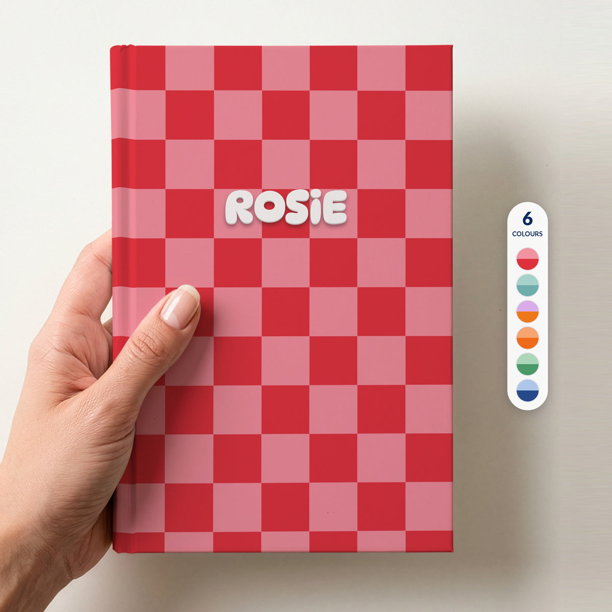 Hand holding a personalised red and pink checkerboard hardcover notebook with the name Rosie in puffy white letters, beside a column of 6 colour swatches showing all 6 colourways
