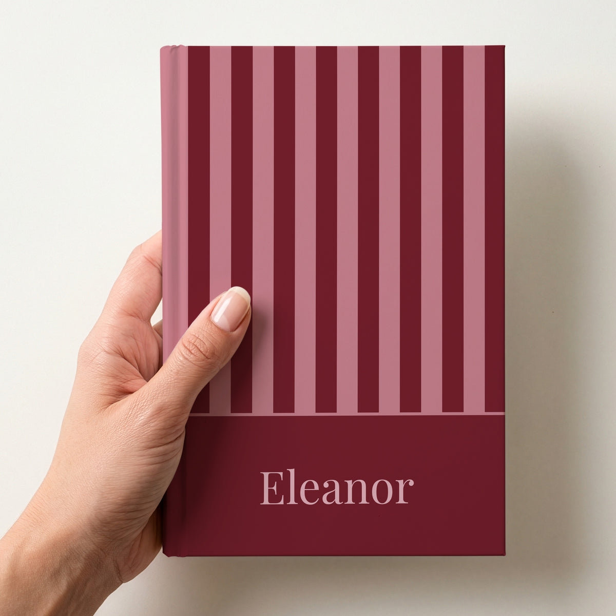 Hand holding a personalised burgundy and pink striped notebook with the name Eleanor in a serif font
