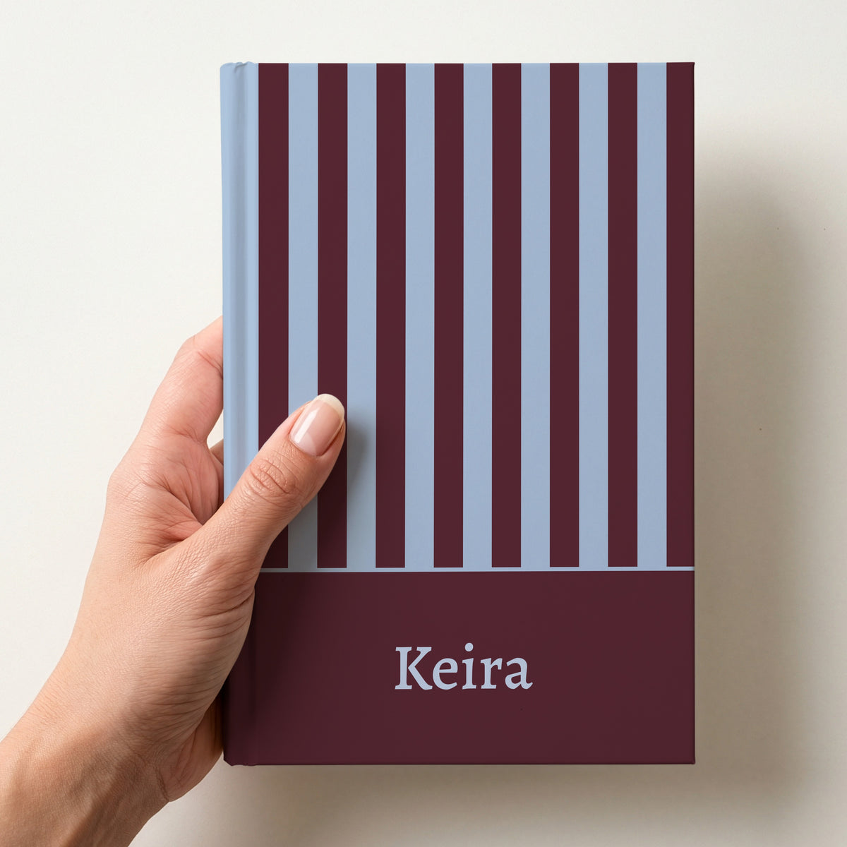 Hand holding a personalised maroon and sky blue striped notebook with the name Keira