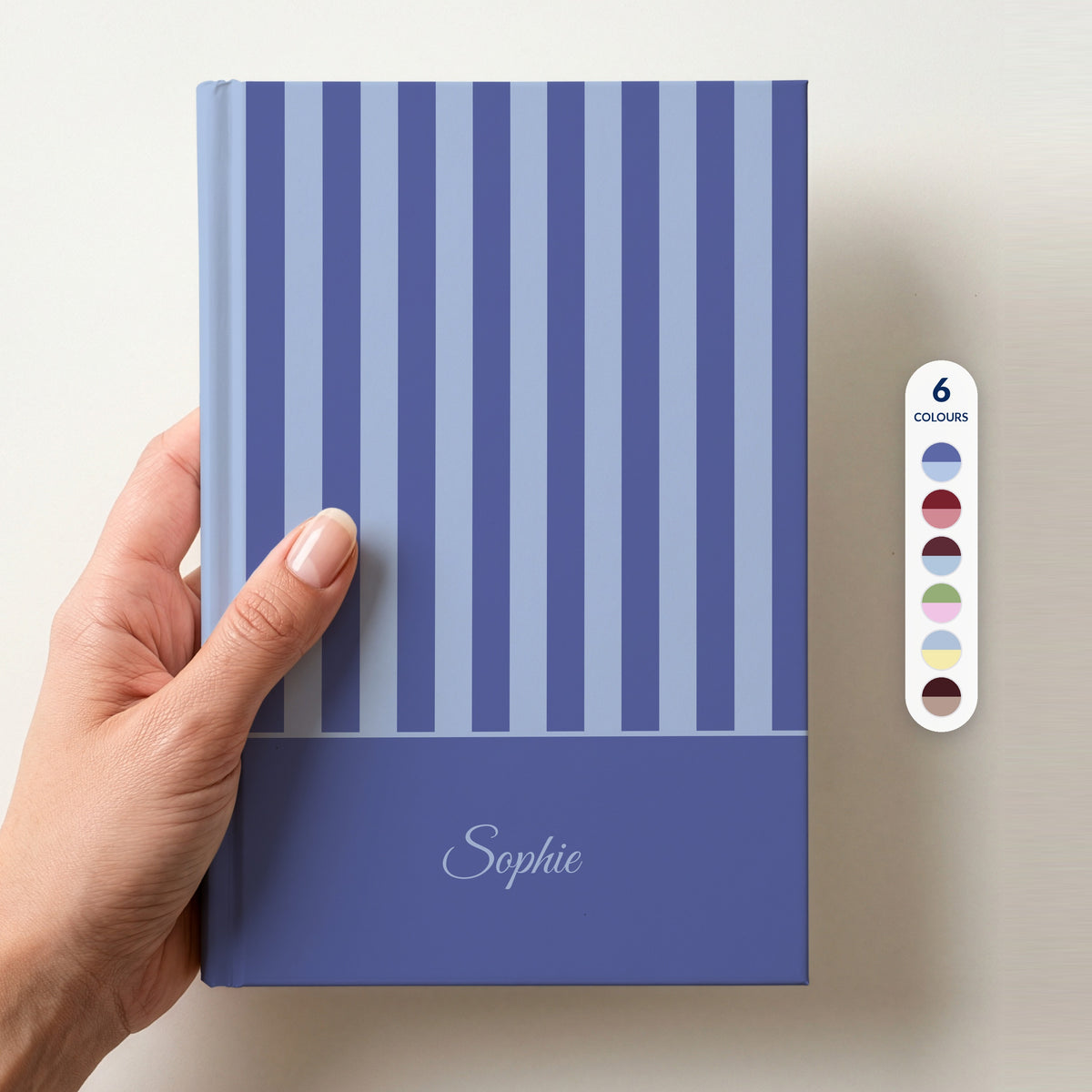 Hand holding a personalised blue striped notebook with a dark blue band and the name Sophie in script, beside a column of 6 colour swatches showing all 6 colourways