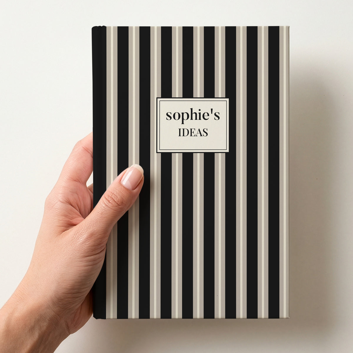 Hand holding a personalised black and stone striped notebook with a label reading sophie's Ideas