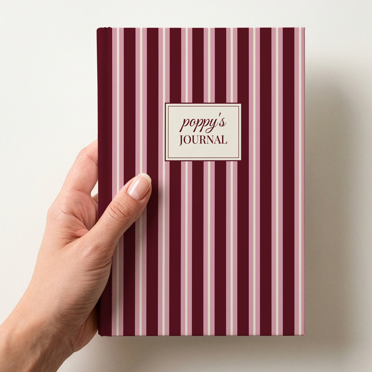 Hand holding a personalised burgundy and pink striped notebook with a label reading poppy's Journal