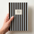 Hand holding a personalised chocolate brown and powder blue striped notebook with a cream label reading olivia's Notes