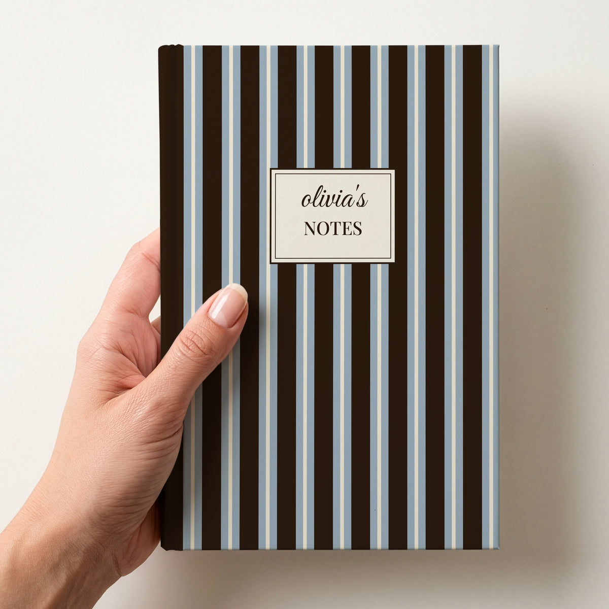 Hand holding a personalised chocolate brown and powder blue striped notebook with a cream label reading olivia's Notes