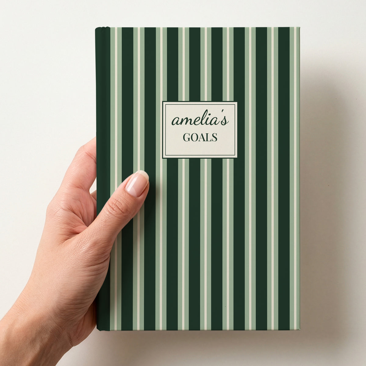 Hand holding a personalised forest green and sage striped notebook with a label reading amelia's Goals in script