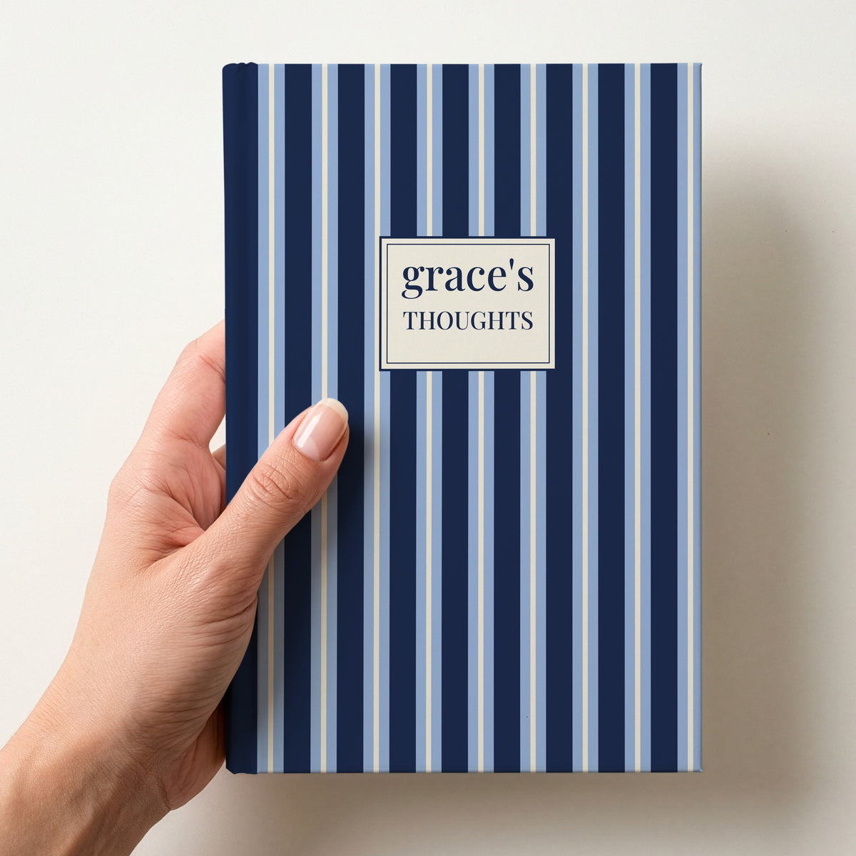 Hand holding a personalised navy and sky blue striped notebook with a label reading grace's Thoughts