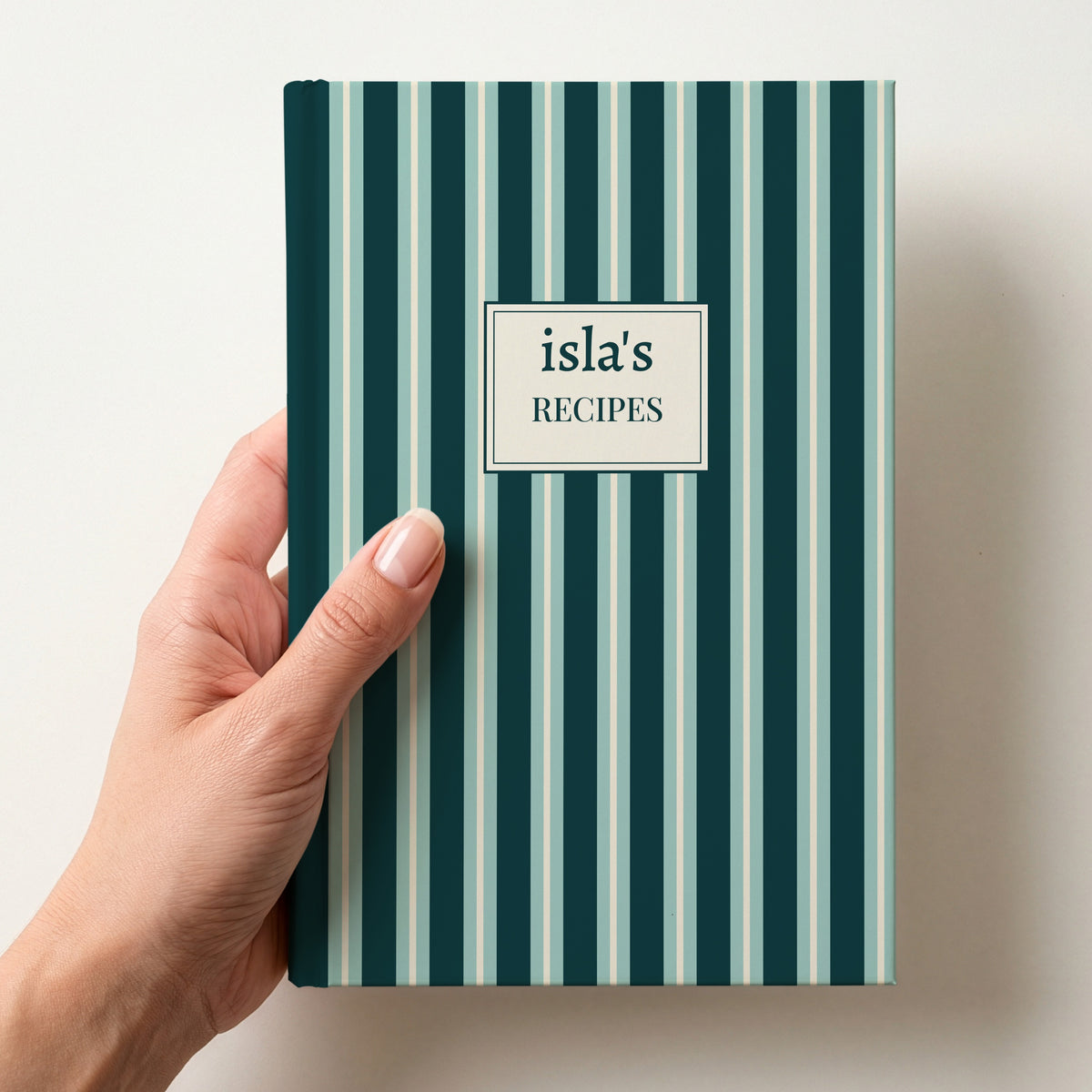 Hand holding a personalised teal and mint striped notebook with a label reading isla's Recipes