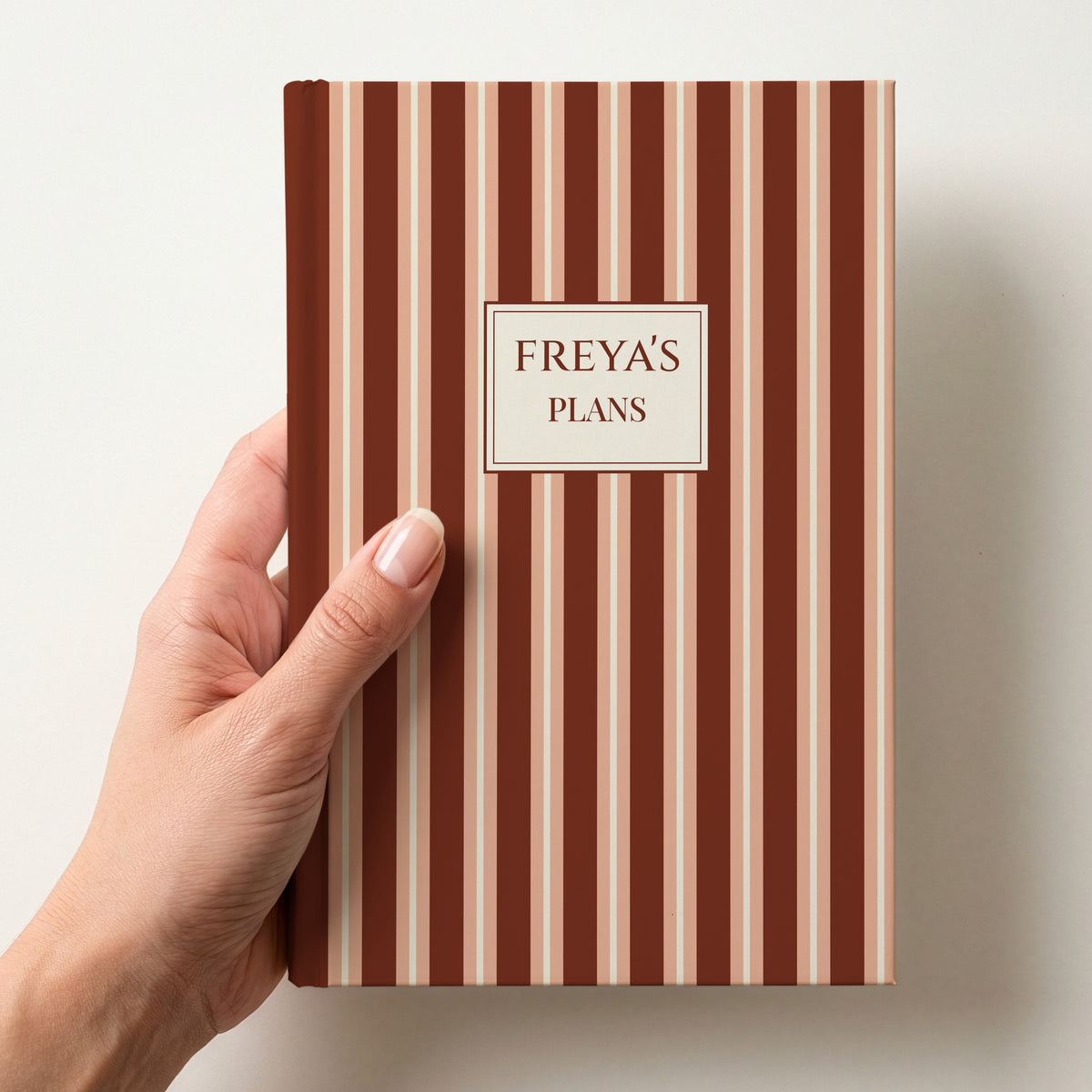Hand holding a personalised terracotta and peach striped notebook with a label reading Freya's Plans in capitals