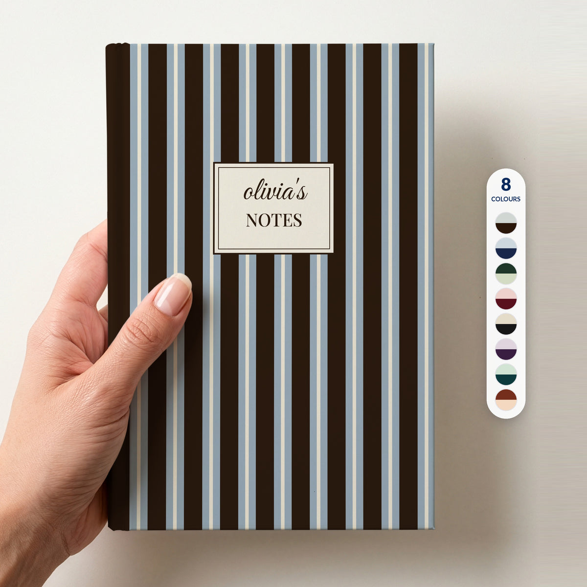 Hand holding a personalised chocolate brown and powder blue striped notebook with a cream label reading olivia's Notes, beside a column of 8 colour swatches showing all 8 colourways