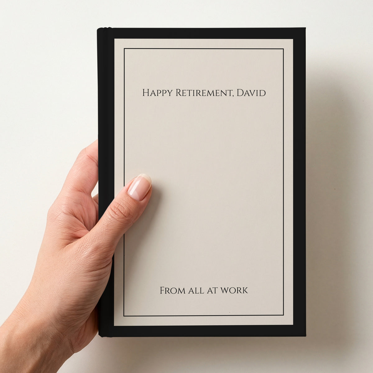 Hand holding a personalised cream notebook with a black border reading Happy Retirement, David and From all at work in capitals
