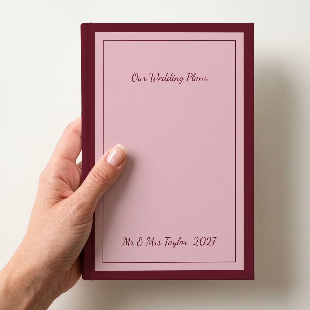 Hand holding a personalised blush pink notebook with a burgundy border reading Our Wedding Plans and Mr & Mrs Taylor 2027