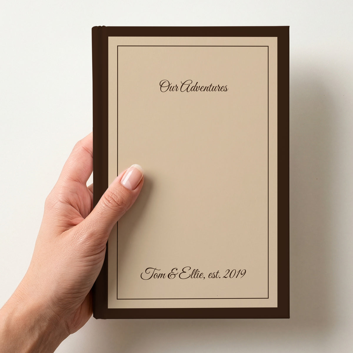 Hand holding a personalised caramel notebook with a chocolate brown border reading Our Adventures and Tom & Ellie, est. 2019 in script