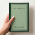 Hand holding a personalised sage green notebook with a forest green border reading Happy 30th Birthday, Sophie and Love, Mum & Dad in script