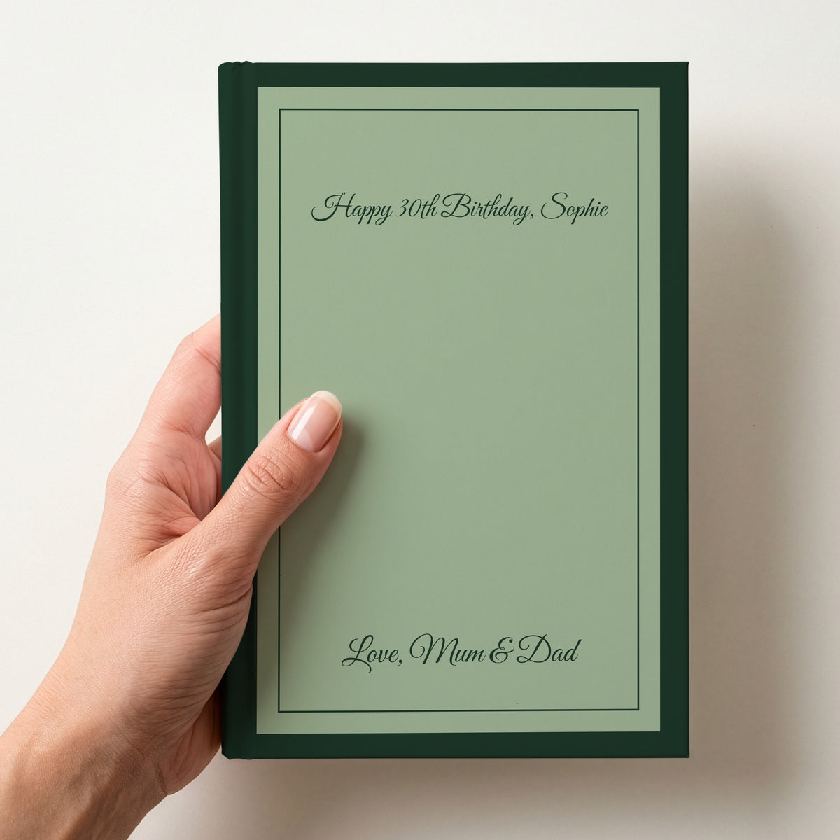 Hand holding a personalised sage green notebook with a forest green border reading Happy 30th Birthday, Sophie and Love, Mum & Dad in script