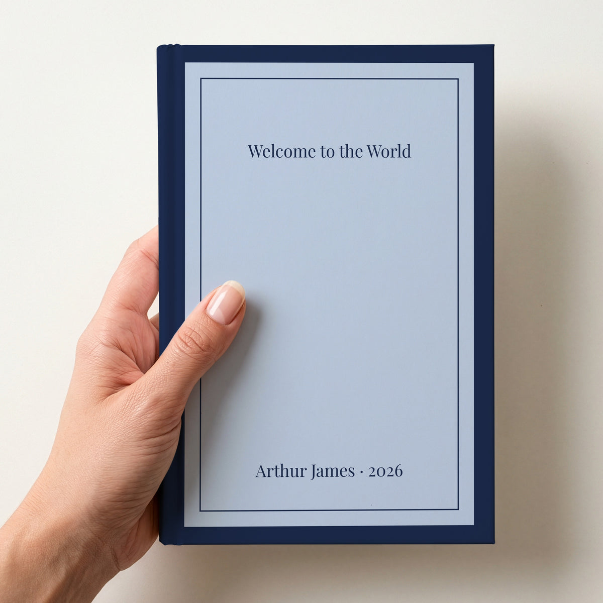 Hand holding a personalised sky blue notebook with a navy border reading Welcome to the World and Arthur James 2026