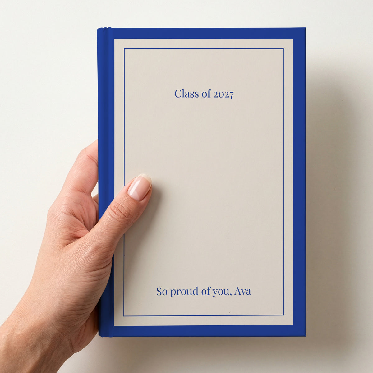 Hand holding a personalised cream notebook with a royal blue border reading Class of 2027 and So proud of you, Ava